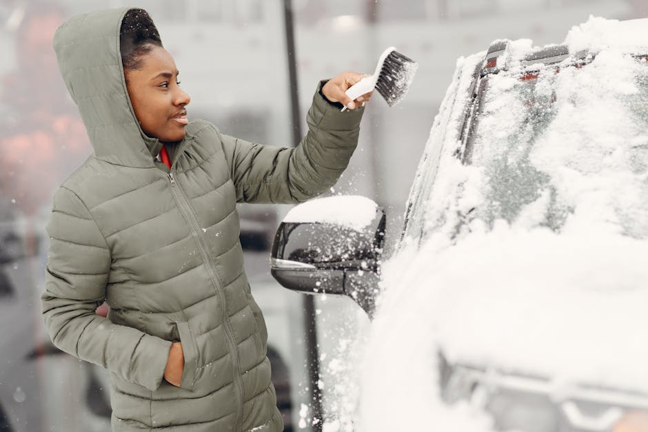 A woman wearing a light grey hooded puffer jacket is standing outdoors in front of a parked vehicle, which is covered in snow. She is holding a black scrub brush and is in the process of clearing snow from the vehicle's windshield. The vehicle appears to be a dark-colored van or large car, positioned on a paved surface next to a building or garage with reflective glass doors. The scene suggests winter conditions, and the woman is preparing the vehicle for transportation or home relocation by removing snow from the windshield, possibly as part of packing and moving preparations. The setting is well-lit, with natural daylight, and the focus is on the woman and the vehicle, supporting themes of vehicle readiness and moving logistics associated with house removals in Hither Green. Man With a Van Hither Green occasionally provides assistance with vehicle preparation during same-day removals or home relocations.