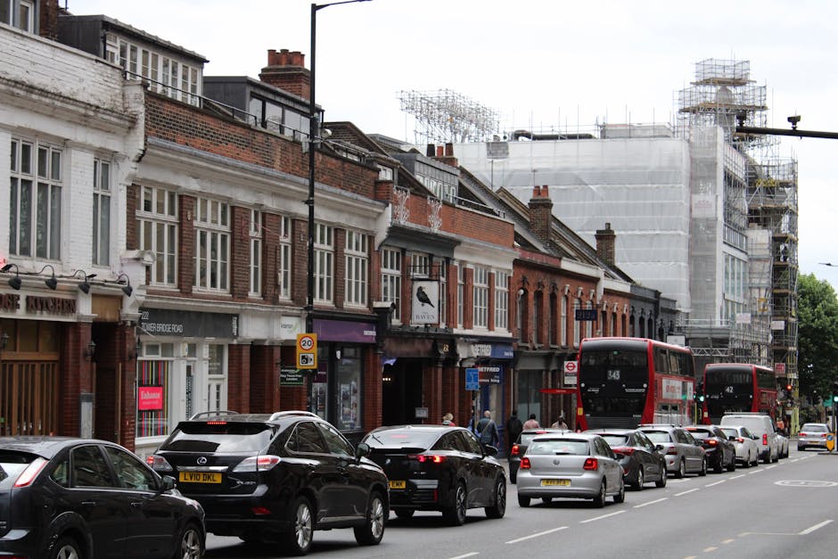 A street scene in Hither Green showing a row of small retail shops and residential buildings with brick facades and white window frames. The street is busy with parked cars lining the curb, including a black SUV, a white hatchback, and several sedans. In addition, two red double-decker buses are visible, one approaching and one passing, indicating a working transport route. To the right, a construction site is covered with white scaffolding and protective sheeting, with structural scaffolding visible above. Pedestrians are seen walking along the sidewalk, some near the shop entrances, and the atmosphere appears typical of urban home relocation or furniture transport activities happening in the area. The lighting is natural and slightly overcast, typical of a cloudy day, supporting the context of potential delays and access issues for house removals in this part of Hither Green, as managed by Man With a Van Hither Green.