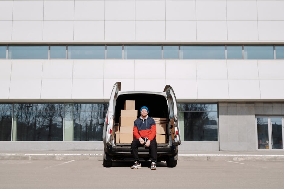 A man in a red and navy jacket and blue beanie sits on the open tailgate of a white commercial van parked outside a modern, multi-storey building with large glass windows. Inside the van's cargo area, several cardboard boxes of different sizes are stacked, some wrapped in plastic and others unwrapped, indicating the process of packing and moving. The man is smiling, and the scene is set on a paved area with no other vehicles or people visible nearby, suggesting a home relocation or furniture transport operation by [COMPANY_NAME]. The background features the building's facade with horizontal window panels, and clear weather provides natural lighting, highlighting the moving equipment such as the van and the neatly arranged boxes ready for loading or unloading, illustrating the vehicle's role in house removals and logistical moving services.