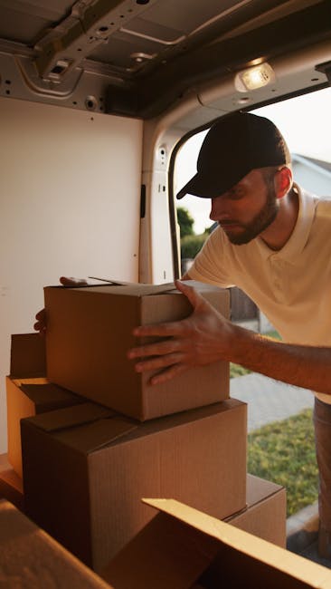 A man wearing a black cap and a light-colored shirt is inside the rear of a moving van, carefully handling several cardboard boxes that are stacked on the vehicle's loading area. The boxes vary in size and are sealed with packing tape, some with flaps open; they are likely filled with household items for a home relocation. The man is lifting or adjusting one box to position it securely on top of another, indicating an organized loading process. The interior of the van shows the metal ceiling and side panels, with a large window on the side allowing natural light to illuminate the scene. Outside the open rear doors, a grassy area and bright sky are visible, suggesting daytime. In the context of house removals and furniture transport, this scene depicts professional packing and loading in progress, consistent with services offered by Man With a Van Hither Green.