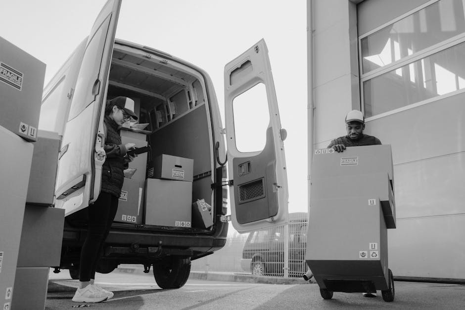 A man in a red and navy jacket and blue beanie sits on the open tailgate of a white commercial van parked outside a modern, multi-storey building with large glass windows. Inside the van's cargo area, several cardboard boxes of different sizes are stacked, some wrapped in plastic and others unwrapped, indicating the process of packing and moving. The man is smiling, and the scene is set on a paved area with no other vehicles or people visible nearby, suggesting a home relocation or furniture transport operation by [COMPANY_NAME]. The background features the building's facade with horizontal window panels, and clear weather provides natural lighting, highlighting the moving equipment such as the van and the neatly arranged boxes ready for loading or unloading, illustrating the vehicle's role in house removals and logistical moving services.