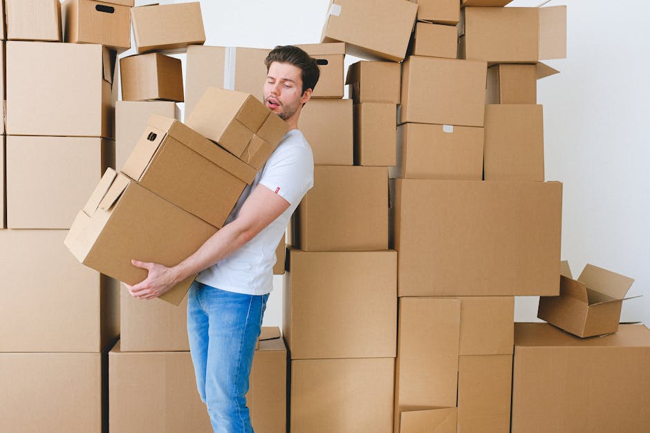 A young man with dark hair and a short beard is inside a room, surrounded by numerous cardboard moving boxes of various sizes, some stacked on top of each other against the wall. He is wearing a white
