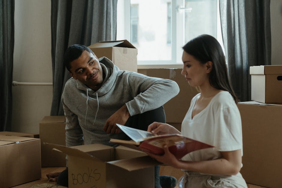 A young man with dark hair and a short beard is inside a room, surrounded by numerous cardboard moving boxes of various sizes, some stacked on top of each other against the wall. He is wearing a white