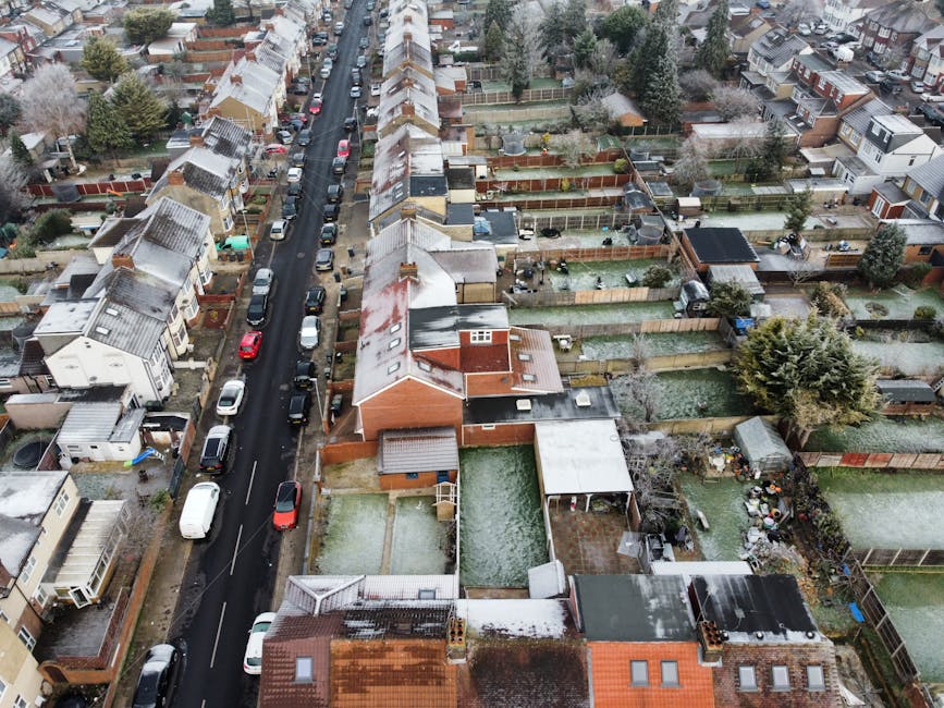 An aerial view of a residential street with closely spaced houses and back gardens in Hither Green, Lewisham. Some roofs and garden fences are dusted with light snow, and various vehicles are parked along the road. Several backyards are visible, featuring fencing, garden furniture, sheds, and decorative plants. The scene shows a typical urban neighbourhood during winter, with a mixture of single- and double-storey houses. In the foreground, a property with a small garden is in the process of a home relocation, with cardboard boxes, plastic packing materials, and furniture covered in moving blankets visible near the property. A man with a van belonging to 'Man With a Van Hither Green' is seen loading furniture and boxes from the driveway onto the vehicle using straps and trolleys, illustrating furniture transport and home moving activities associated with official Lewisham Council removal permits for Hither Green residents. The overall environment depicts a typical moving day within a well-established residential area, emphasizing the logistics of packing and transporting household items during house removals.