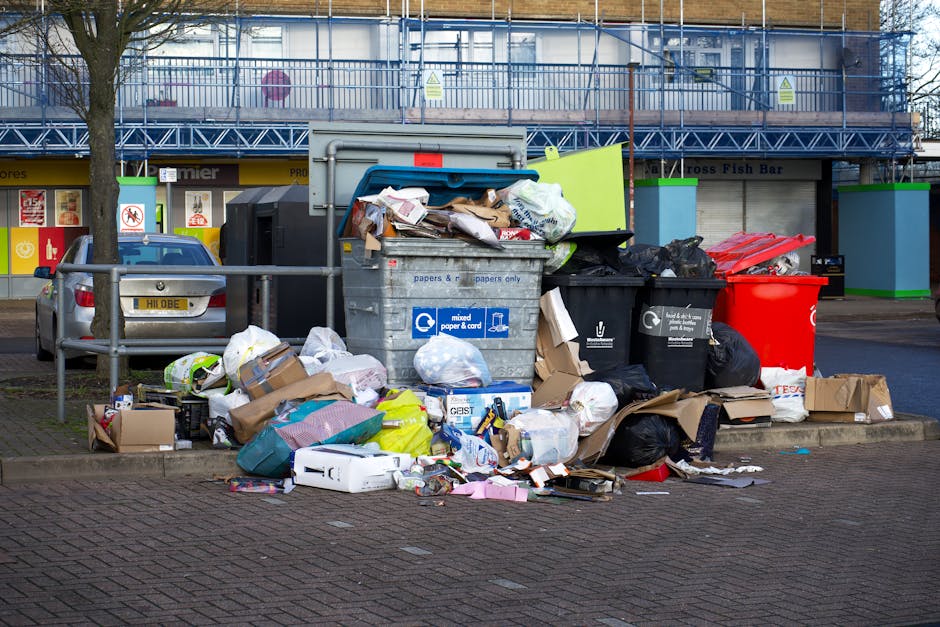 An outdoor scene showing a collection of overflowing recycling and waste bins on a paved pavement next to a parking area. The collection includes a large grey mixed paper and cardboard recycling bin with its lid open, revealing crumpled paper, cardboard boxes, and plastic waste spilling out. Surrounding this bin are several black black rubbish bags, a bright red waste bin, and a black bin with a white recycling symbol, all filled with assorted trash. Cardboard boxes, some flattened and others intact, are scattered on the pavement, along with various plastic and paper packaging. A green trolley and a few smaller loose items, such as paper rolls and discarded packaging, are also visible nearby. Behind the bins, a metal railing separates the waste area from parked cars, including a grey vehicle with a visible license plate, and a large building under construction with scaffolding and protective netting. The scene captures the typical logistics of waste disposal that may be seen during a home or office relocation process, with Man With a Van Hither Green often assisting with such moving and clearing tasks.