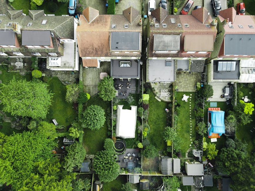 An aerial view of a residential street with closely spaced houses and back gardens in Hither Green, Lewisham. Some roofs and garden fences are dusted with light snow, and various vehicles are parked along the road. Several backyards are visible, featuring fencing, garden furniture, sheds, and decorative plants. The scene shows a typical urban neighbourhood during winter, with a mixture of single- and double-storey houses. In the foreground, a property with a small garden is in the process of a home relocation, with cardboard boxes, plastic packing materials, and furniture covered in moving blankets visible near the property. A man with a van belonging to 'Man With a Van Hither Green' is seen loading furniture and boxes from the driveway onto the vehicle using straps and trolleys, illustrating furniture transport and home moving activities associated with official Lewisham Council removal permits for Hither Green residents. The overall environment depicts a typical moving day within a well-established residential area, emphasizing the logistics of packing and transporting household items during house removals.