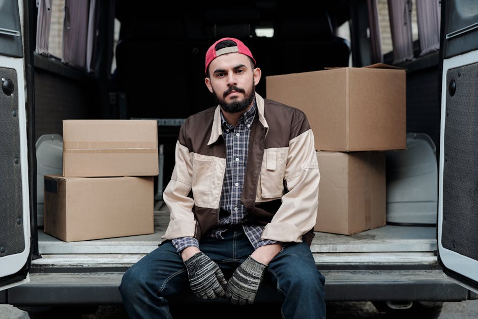 A young man with a beard, wearing a red cap backwards, a brown and beige jacket, a checked shirt, and striped gloves, sits on the edge of an open van with the rear doors wide open. Inside the vehicle, there are several cardboard boxes of varying sizes, some stacked on top of each other, prepared for home relocation. The boxes are secured with packing tape, and the van's interior is visible with dark seating and a clean cargo area. The scene is well-lit, showing the outdoor environment where packing and furniture transport are in progress as part of the loading process. The image reflects professional removal services by Man With a Van Hither Green, capturing a moment during the logistics of packing and loading for a house move, highlighting the importance of careful handling of boxes and furniture during relocation.