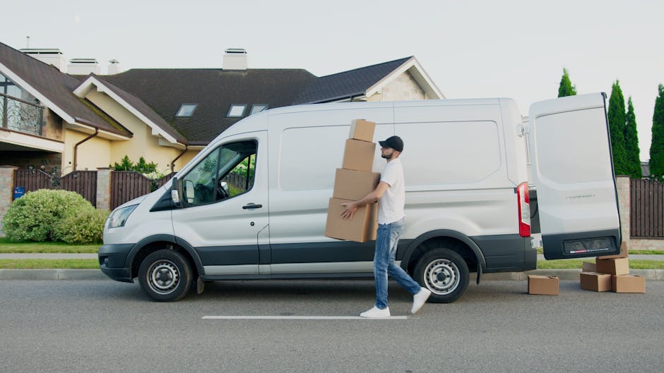 A man wearing casual clothing, including a black cap, white t-shirt, and jeans, is standing beside a white removal van parked on a residential street during daytime. He is carrying three cardboard boxes stacked one on top of the other, with additional boxes on the pavement nearby. The van's side door is open, indicating an active loading process for home relocation or furniture transport. The background features a modern, two-storey house with a pitched roof, skylights, a covered porch, and a fence with landscaping including bushes and small trees. The scene is well-lit with natural daylight, and the overall environment suggests a professional moving operation by Man With a Van Hither Green, involved in packing and moving services.