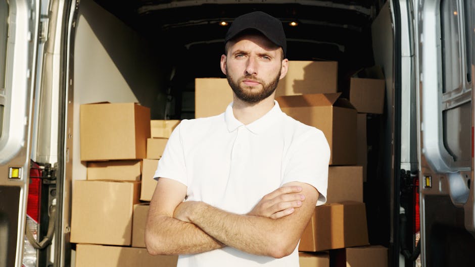 A young man with a beard, wearing a red cap backwards, a brown and beige jacket, a checked shirt, and striped gloves, sits on the edge of an open van with the rear doors wide open. Inside the vehicle, there are several cardboard boxes of varying sizes, some stacked on top of each other, prepared for home relocation. The boxes are secured with packing tape, and the van's interior is visible with dark seating and a clean cargo area. The scene is well-lit, showing the outdoor environment where packing and furniture transport are in progress as part of the loading process. The image reflects professional removal services by Man With a Van Hither Green, capturing a moment during the logistics of packing and loading for a house move, highlighting the importance of careful handling of boxes and furniture during relocation.