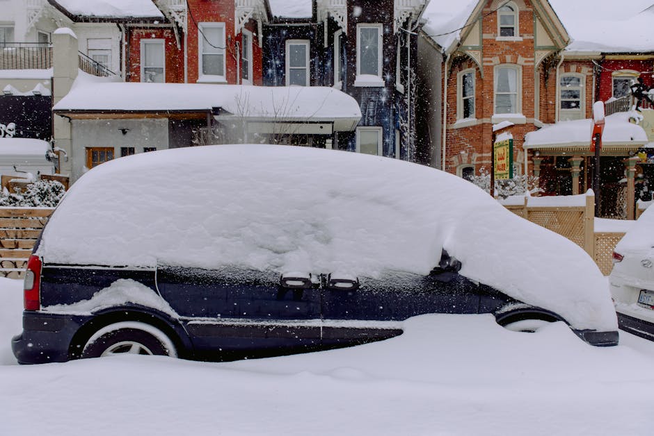 A black van parked on a snow-covered street in a residential area, with its windows and roof heavily encrusted with snow during a winter snowfall; the vehicle is situated close to the pavement and appears to be part of a home relocation or moving process. In the background, there are multi-storey houses with detailed brickwork and bay windows, also snow-covered, indicating cold weather and recent snowfall. Snowflakes are falling, creating a wintry scene, and the surrounding environment includes a wooden fence and parked cars. The scene suggests that Man With a Van Hither Green is preparing for or has just completed a furniture transport or household packing and moving service in this residential area, with snow and winter weather conditions impacting the logistics and timing of the home relocation.