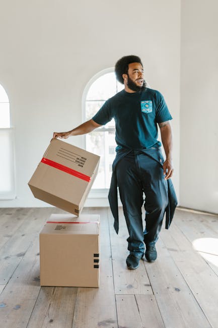 A man with a dark complexion, curly black hair, and a beard is standing inside a bright, empty room with white walls and wooden flooring. He is wearing a blue T-shirt with a light blue geometric pattern on the chest, dark trousers, and has a dark jacket tied around his waist. He is holding a cardboard box with red tape in his right hand, preparing to place it on top of another box on the floor. Behind him, there is a large arched window allowing natural light to illuminate the space. The room appears to be in the process of home relocation, with the man involved in the packing and furniture transport activities typical of professional house removals. The scene reflects the logistical steps of loading boxes during a house move, as part of a comprehensive moving service provided by Man With a Van Hither Green, aligning with the detailed move planning and packing process in the home relocation industry.