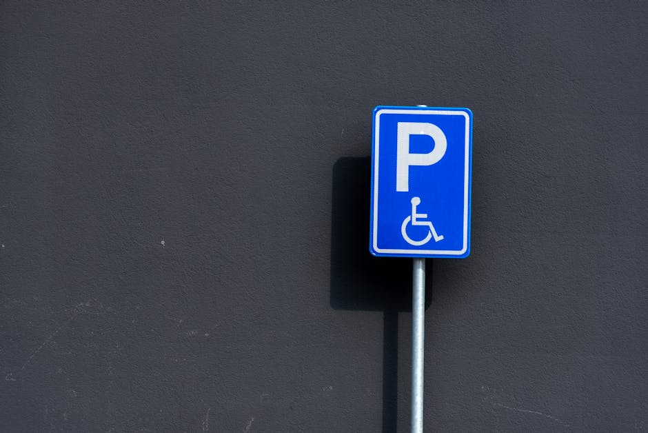 A blue and white parking sign indicating accessible parking for disabled persons, mounted on a metallic pole against a dark grey wall. The sign features a large white letter 'P' and a wheelchair symbol below it, signifying designated parking for individuals with mobility impairments. The sign is positioned on a paved area, likely outside a property, and is captured in natural daylight, with a shadow cast on the wall behind it. This image relates to parking regulations for house removals and moving services, such as those provided by Man With a Van Hither Green, assisting with parking permits in Lewisham borough for home relocation and furniture transport.