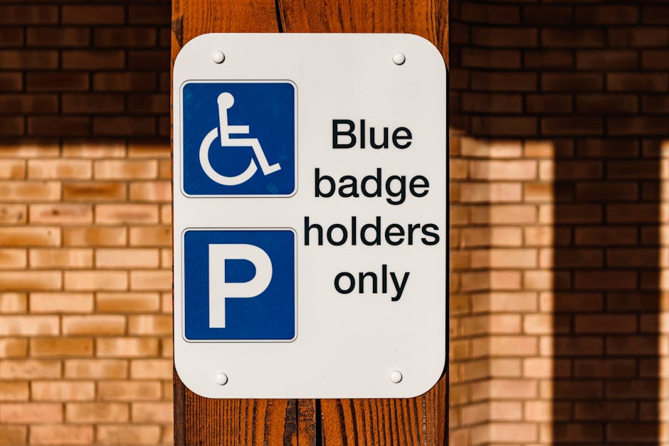 A rectangular white sign attached to a wooden post outdoors, featuring a blue wheelchair symbol at the top left corner, indicating accessibility, with the text 'Blue badge holders only' beside it, and a large blue parking symbol 'P' below. The sign is positioned in front of a brick wall illuminated by sunlight, suggesting it is near a parking area. This sign relates to parking regulations for individuals with blue badges, relevant to house removals and loading processes. Occasionally, [COMPANY_NAME], such as Man With a Van Hither Green, may assist with moving logistics and furniture transportation in compliance with local parking rules, including those indicated by this sign.